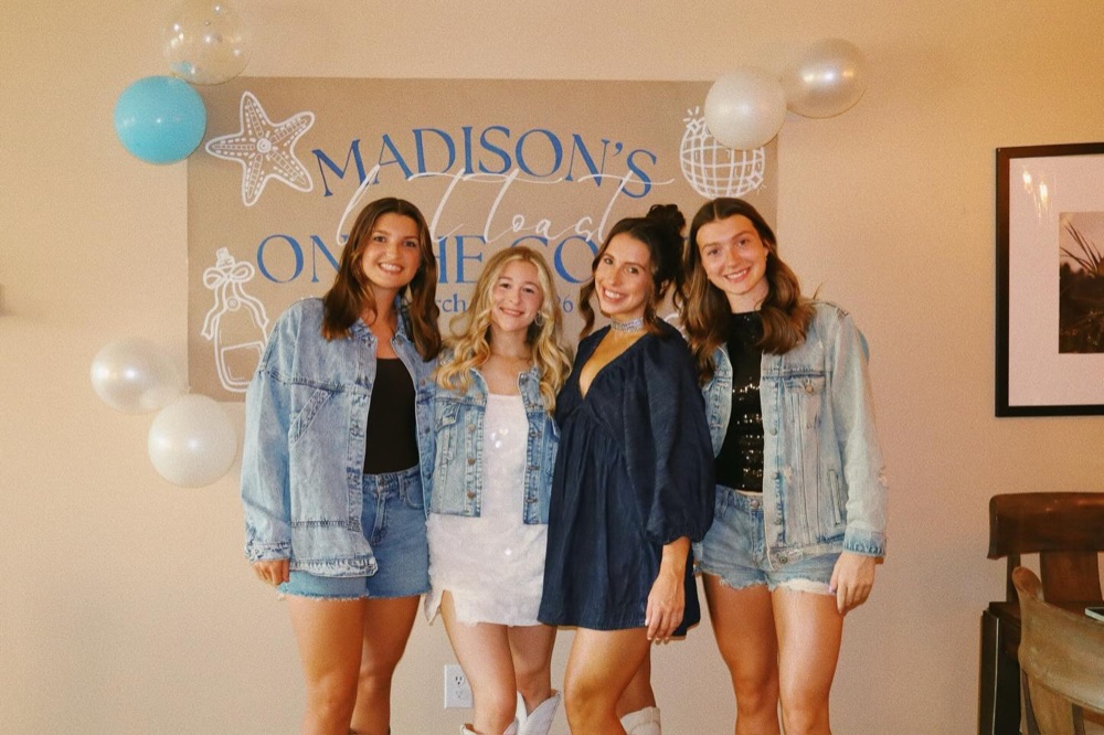 Four friends in denim jackets in front of the Madison's Last Toast on the Coast bachelorette banner