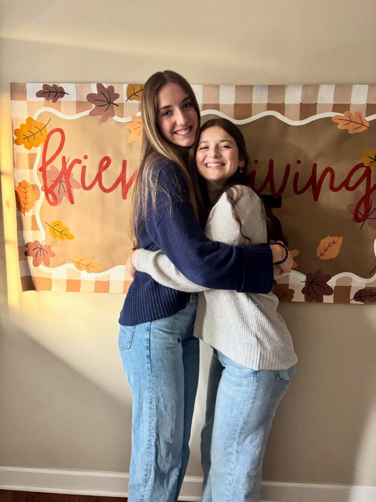Two friends hugging in front of a fall-themed Friendsgiving banner